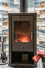 Modern Wood Burning Stove With Accessories And Red Gloves Next To It. Wood Burning Stove With Orange Yellow Flames.