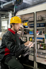 Poster - Contemporary young engineer of factory in workwear and hardhat using tablet