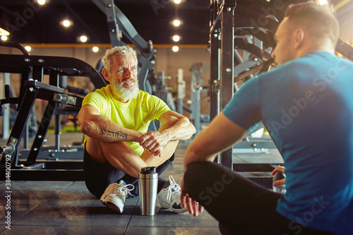 Resting after training. Athletic middle aged man discussing something with fitness instructor or personal trainer while sitting together on the floor at gym