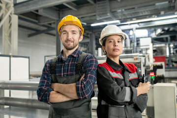 Poster - Two young confident workers in helmets and workwear standing in large workshop