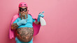 Superhero ready for spring cleaning. Plump bearded housekeeper in costume poses with detergent and indicates away on blank space over pink background. Domestic service. Professional clean up