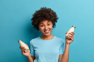 Wall Mural - Vegan food and nutrition concept. Cheerful dark skinned woman with curly hair holds bottle of fresh almond milk shows healthy vegetarian product smiles happily isolated on blue studio background