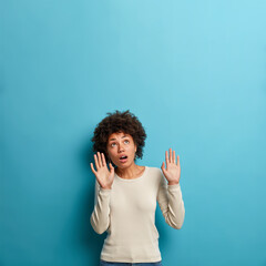 Wall Mural - Vertical shot of fearful young Afro American woman keeps palms forward and looks with fright above on ceiling tries to defence herself from invisible danger opens mouth dressed in casual jumper