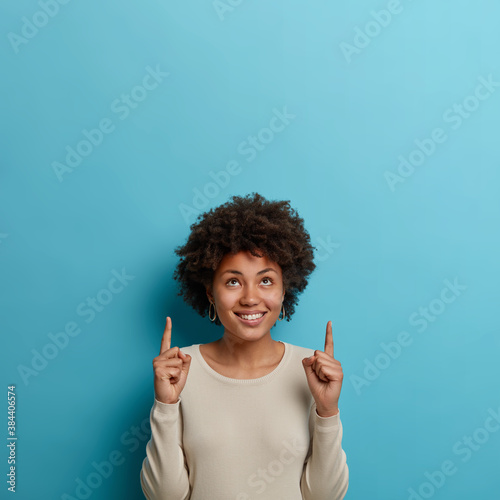 Joyful dark skinned African American woman with curly hair points above over head indicates something on blank blue studio wall has happy smile dressed casually. People and advertising concept