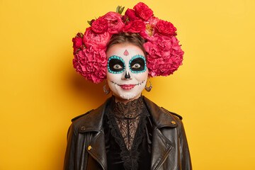 Wall Mural - Cheerful female with artistic halloween makeup wears traditional attire for mexican day of death to honor dead relatives has spooky image poses against yellow background. Costume masquerade.