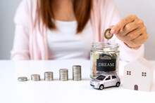 Young Asian Woman's Hand Putting Coin In Glass Jar With Model Home And Car On White Table Background. Saving, Collect Money For Future, Investment Concept. Close Up