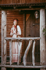 A beautiful Slavic girl with long blonde hair and brown eyes in a white and red embroidered suit stands on the porch of a house.Traditional clothing of the Ukrainian region.