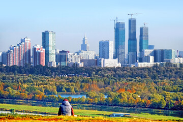 Wall Mural - Modern moscow city skyline landmark at autumn. Person sitting on hill against cityscape background. Colorful residential buildings. Golden autumn urban landscape