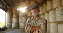 Portrait Of Caucasian Old Gray-haired Male Farmer In Cap Standing At Hay Stocks In Farm And Looking At Camera In Sunlight. Grandfather Worker At Village. Countryside Concept. Senior Man.
