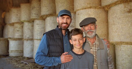 Wall Mural - Three male generations in shed at hay stocks. Caucasian grandfather, father and son smiling to camera and standing in barn. Farming concept. Countryside. Old man farmer with son and grandson.