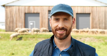 Wall Mural - Portrait of Caucasian young smiled happy man with beard posing to camera with hands on sides and smiling. Handsome male farmer standing at field pasture with sheep grazing on background.