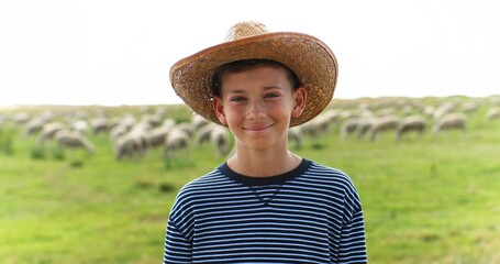 Wall Mural - Portrait of Caucasian teen small happy boy in hat standing in green field in summer and smiling to camera. Sheep flock grazing on background. Little teenager shepherd. Vacations at countryside farm.