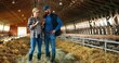 © VAKSMANV - Caucasian couple farmers walking in barn with sheep flock, talking and using tablet device. Shepherds strolling in stable with livestock and having conversation. Man and woman watching video on gadget