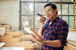 © DragonImages - Smiling Asian carpenter looking at wooden brick in his hand when recording voice message to customer