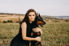 Young Woman Sitting On Green Grass, Hugging A Doberman Breed Dog, Outdoors.