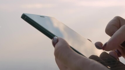Close up view of woman hand using smartphone device with touchscreen display against clear sky - scrolling and touching. Leisure time, entertainment, communication, technology concept