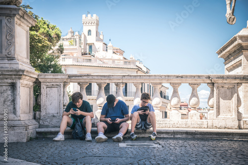 Group of teenagers playing with smartphone sitting in a historic square in Rome. Young boys using smart devices outdoors to find tourist map during holiday. Tech, communication, youth, tourism concept