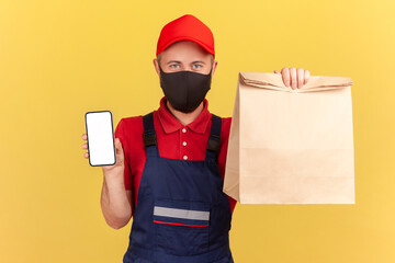 Deliveryman in uniform and protective mask holding and showing paper bag and smartphone with empty screen, application for food delivery, advertising. Indoor studio shot isolated on yellow background