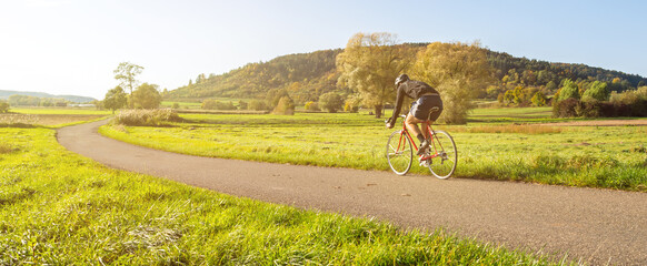 Wall Mural - Panorama shot of cyclist on a racing bike in scenic rural autumn landscape during beautiful afternoon light