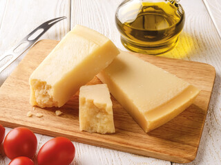 Wall Mural - Wedge of italian hard cheese Parmesan or Grana Padano on a brown wood cutting boad near small red tomatoes, olive oil and cheese knife over white wood table. High key image.