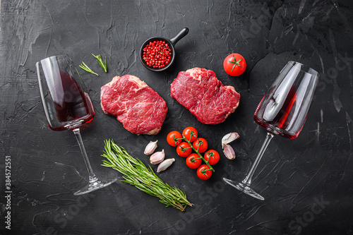 Raw rump steaks from organic beef meat cuts near red wine glasses with rosemary, garlic and spices over black textured  background, top view.