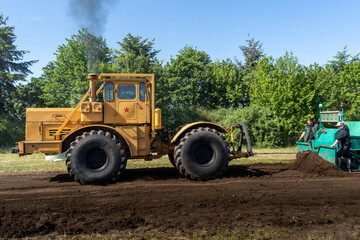 Yellow tractor in a field with dark steam coming out ouf exhaust, pulling a heavy weight