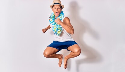 Young handsome hispanic tourist man on vacation wearing summer clothes and hawaiian lei smiling happy. Jumping with smile on face celebrating with fists up over isolated white background