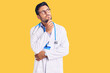 Young hispanic man wearing doctor uniform and stethoscope with hand on chin thinking about question, pensive expression. smiling with thoughtful face. doubt concept.