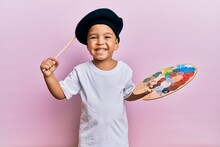 Adorable Latin Toddler Smiling Happy Wearing Artist Style Using Paintbrush And Palette Over Isolated Pink Background.