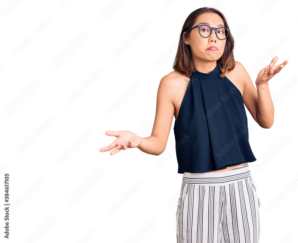 Young beautiful chinese girl wearing casual clothes and glasses clueless and confused expression with arms and hands raised. doubt concept.