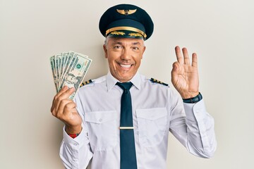 Wall Mural - Handsome middle age mature man wearing airplane pilot uniform holding dollars doing ok sign with fingers, smiling friendly gesturing excellent symbol