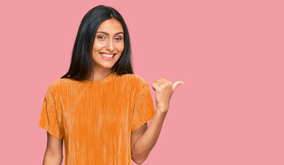 Young brunette arab woman wearing casual clothes smiling with happy face looking and pointing to the side with thumb up.