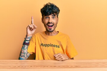 Young hispanic man wearing tshirt with happiness word message sitting on the table pointing finger up with successful idea. exited and happy. number one.