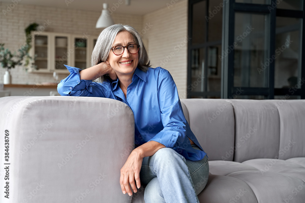 Happy relaxed mature old adult woman wearing glasses resting sitting on couch at home. Smiling mid age grey-haired elegant senior lady relaxing on comfortable sofa looking at camera. Portrait - obrazy, fototapety, plakaty 