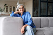 Happy relaxed mature old adult woman wearing glasses resting sitting on couch at home. Smiling mid age grey-haired elegant senior lady relaxing on comfortable sofa looking at camera. Portrait