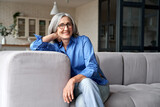 Fototapeta  - Happy relaxed mature old adult woman wearing glasses resting sitting on couch at home. Smiling mid age grey-haired elegant senior lady relaxing on comfortable sofa looking at camera. Portrait