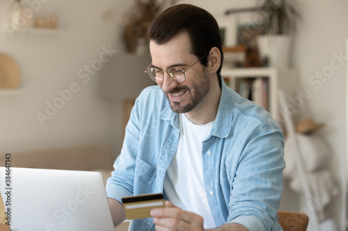 Happy young man in eyeglasses enjoying shopping in popular internet store on laptop, making purchase of goods or services using information from plastic banking credit card, secure payment concept.