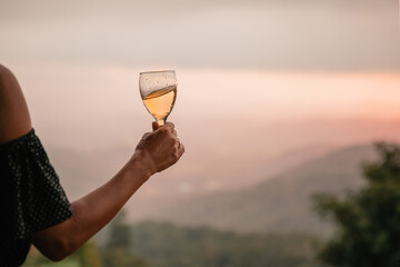 Wall Mural - Woman's hand holding glass of white wine across beautiful mountains landscape and sunset