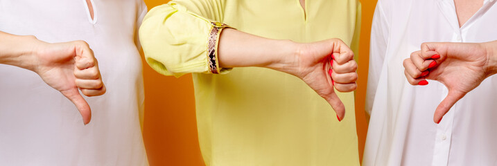 Wall Mural - Rejecting sign. Wrong choice. Criticizing women. Bad idea. Group portrait of three ladies in white yellow blouse disapproving offer with thumbs down dislike hand gesture isolated on orange background.