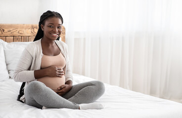 Wall Mural - Pregnancy Time. Happy Black Pregnant Woman Sitting On Bed Touching Belly