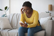 © Prostock-studio - Worried black lady looking at smartphone screen, sitting on couch at home
