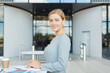 © Seventyfour - Waist up portrait of elegant blonde businesswoman smiling at camera while standing by cafe table in airport, copy space