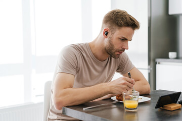 Young man in earphones looking at cellphone while having breakfast at home