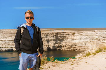 Attractive young man stands on a high cliff overlooking the rocky cliffs arches on the beach and turquoise sea water on the coast