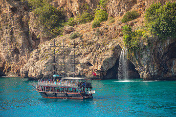 Wall Mural - View of waterfall at Mediterranean seacoast cliff and beach near Antalya and big tour boat resembling a pirate sailing ship from the past, Antalya, Turkey.