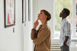 © Seventyfour - Side view portrait of elegant young woman looking at paintings while exploring modern art gallery exhibition, copy space