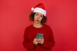 © Jihan - Portrait of a confused Caucasian woman wearing Christmas hat holding mobile phone and shrugging shoulders and frowning face over isolated over red background