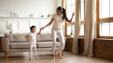 Overjoyed Young Asian Mother And Little Daughter Dancing At Home, Smiling Mum And Adorable Toddler Girl Child Moving To Music, Holding Hands, Standing In Living Room, Family Enjoying Funny Activity