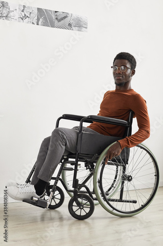 Vertical full length portrait of young African-American man using wheelchair and looking at camera while exploring modern art gallery exhibition