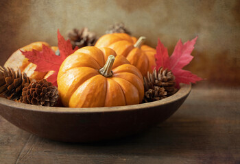 Original textured still life photograph of a wooden bowl of orange mini pumpkins and little pinecones with red maple leaves on brown